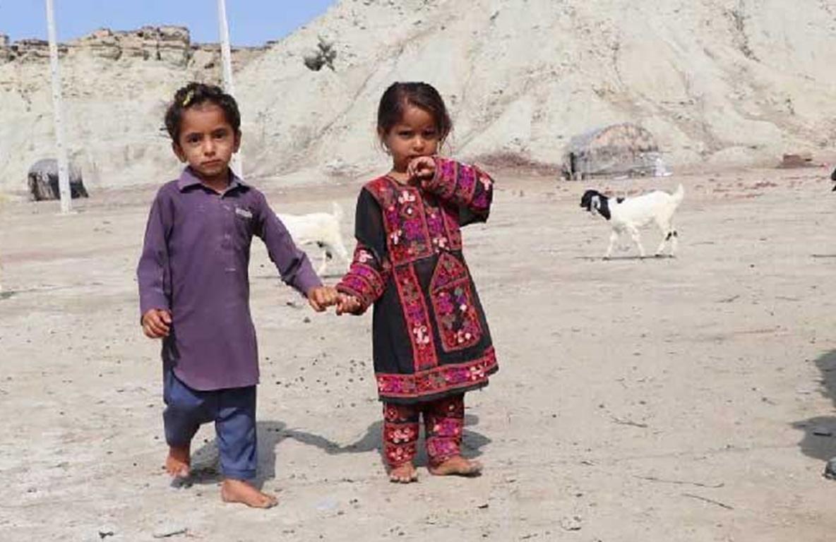 Two young children holding hands and walking in a sandy area with goats in the background.